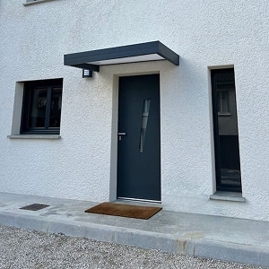 Porte d'entrée anthracite et marquise pour maison moderne Porte d'entrée anthracite avec marquise plate et éclairage intégré sur façade blanche de maison moderne.