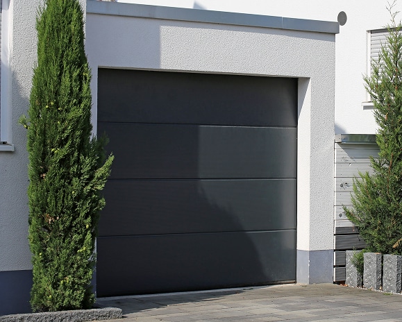Porte de Garage Sectionnelle Grise et Moderne pour Maison Porte de garage sectionnelle gris anthracite moderne installée sur une façade de maison blanche avec arbustes.