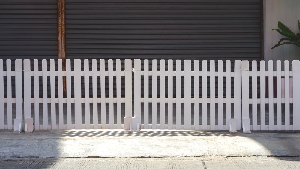 Clôture Blanche en Bois devant Rideau Métallique Gris Clôture en bois blanc devant un rideau métallique gris avec des ombres nettes sur le sol ensoleillé.