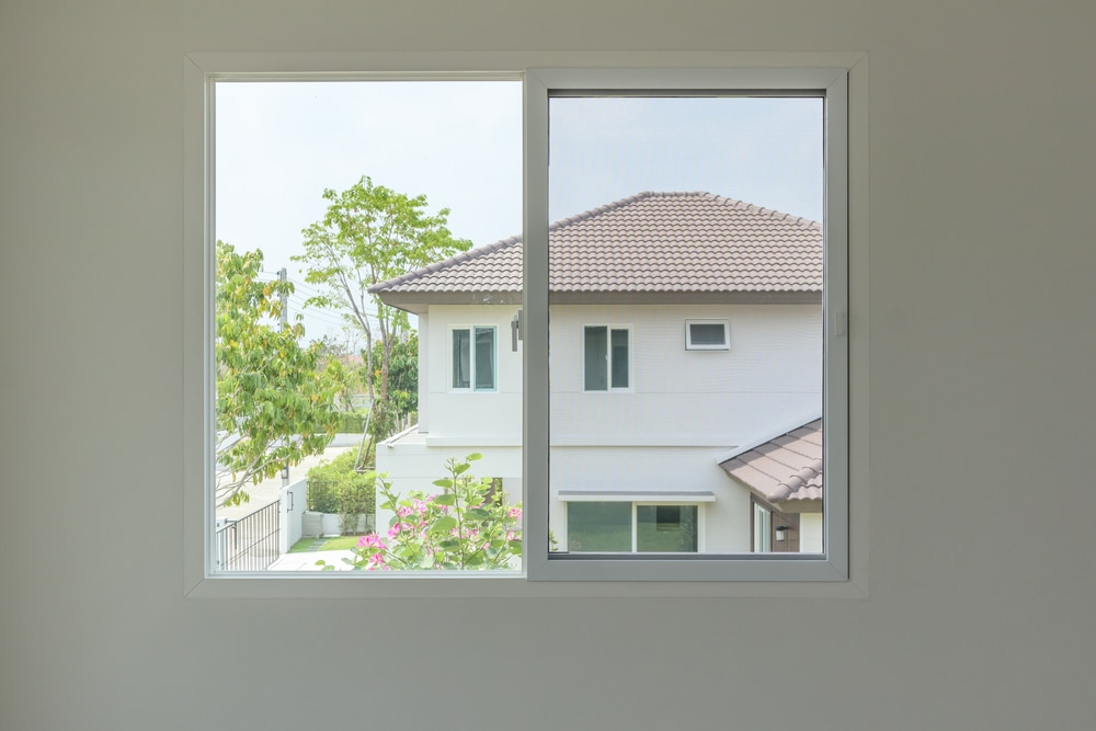 Fenêtre coulissante avec vue sur maison moderne et jardin Vue d'une fenêtre blanche coulissante sur une maison moderne avec jardin fleuri et toit en tuiles brunes.