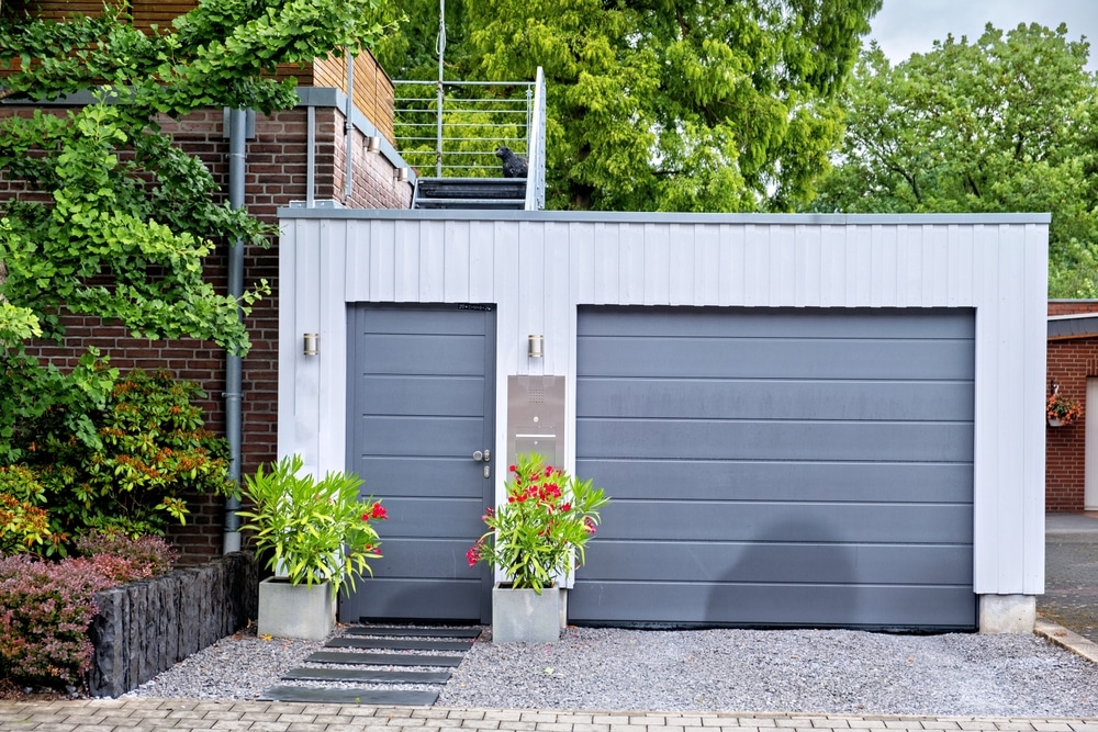 Garage Moderne Blanc et Gris avec Porte Sectionnelle Garage moderne à bardage blanc, porte sectionnelle grise et entrée latérale avec plantes fleuries sur gravier.