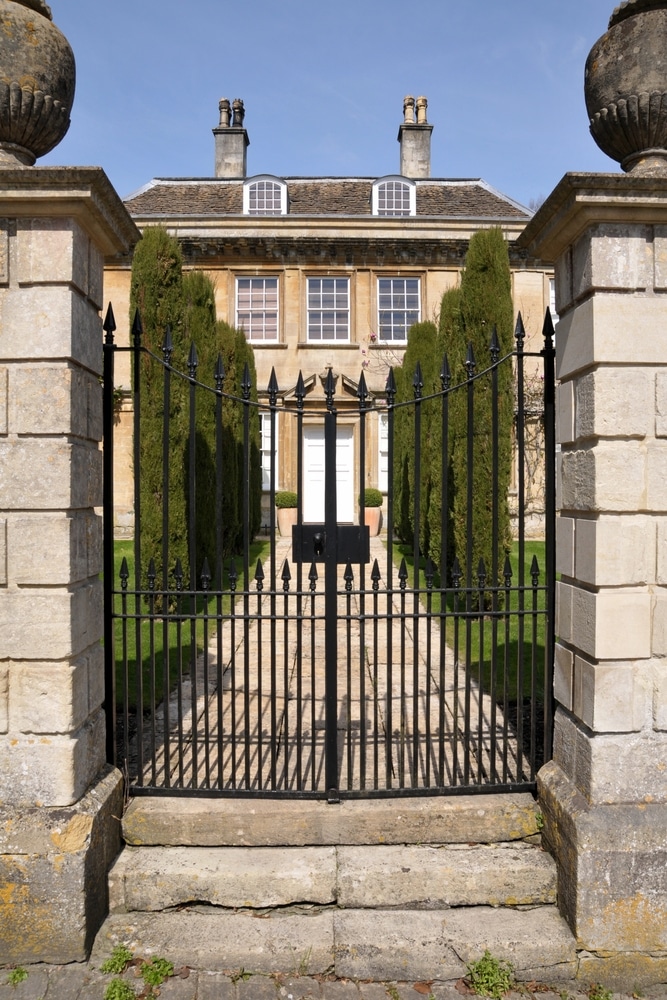 Manoir en pierre et portail en fer forgé classique Manoir en pierre derrière un portail en fer forgé noir, allée pavée et cyprès sous un ciel bleu dégagé.