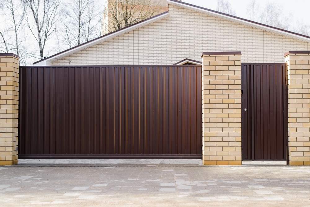 Portail coulissant marron et piliers en briques jaunes Portail coulissant et portillon en métal marron entre des piliers de briques jaunes devant une maison moderne.