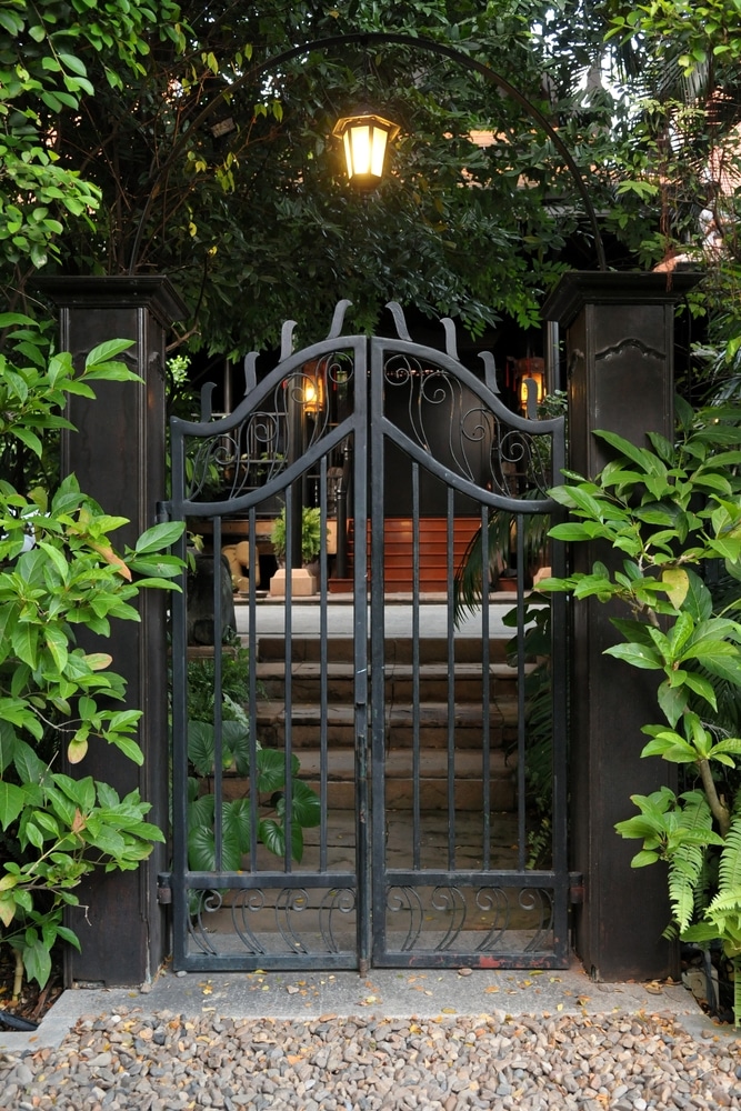 Portail en Fer Forgé et Lanterne pour Entrée de Jardin Portail noir en fer forgé devant un jardin verdoyant avec une lanterne suspendue sous une arche métallique.
