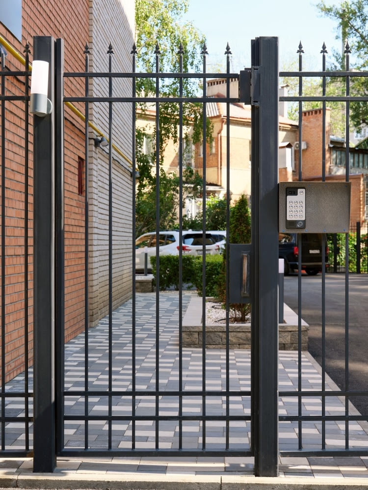 Portail en Fer Noir avec Digicode pour Accès Sécurisé Portail noir en fer avec digicode ouvrant sur une cour pavée et des bâtiments résidentiels en briques.
