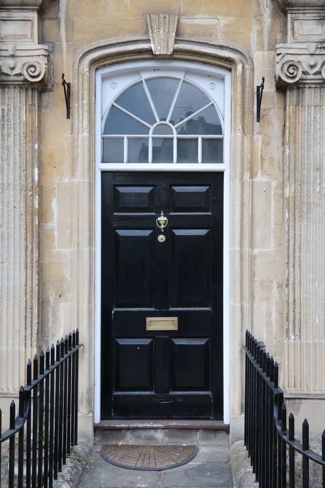Porte d'entrée noire géorgienne avec imposte et pierre Porte noire de style géorgien avec heurtoir en laiton, imposte en demi-lune et façade en pierre entourée de grilles.