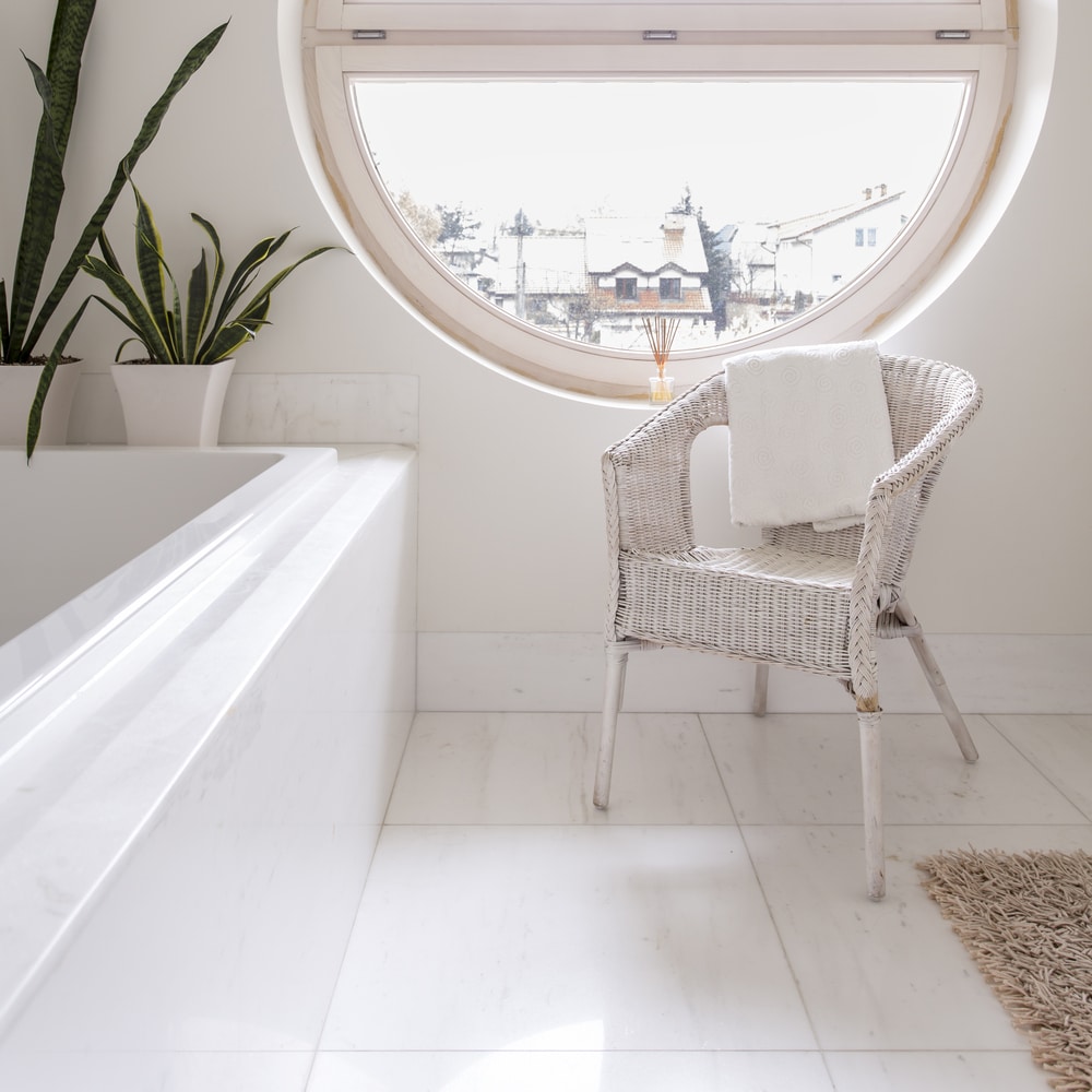 Salle de Bain Lumineuse avec Fenêtre Ronde et Déco Zen Salle de bain zen avec baignoire, fenêtre ronde, plantes et chaise en osier blanc sur sol en marbre.