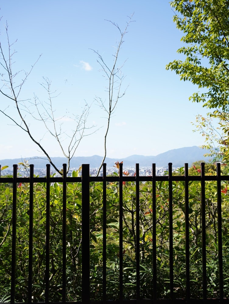 Vue Panoramique sur la Ville et les Montagnes à l'Horizon Vue panoramique d'une ville et de montagnes à travers une clôture noire et des buissons sous un ciel bleu.