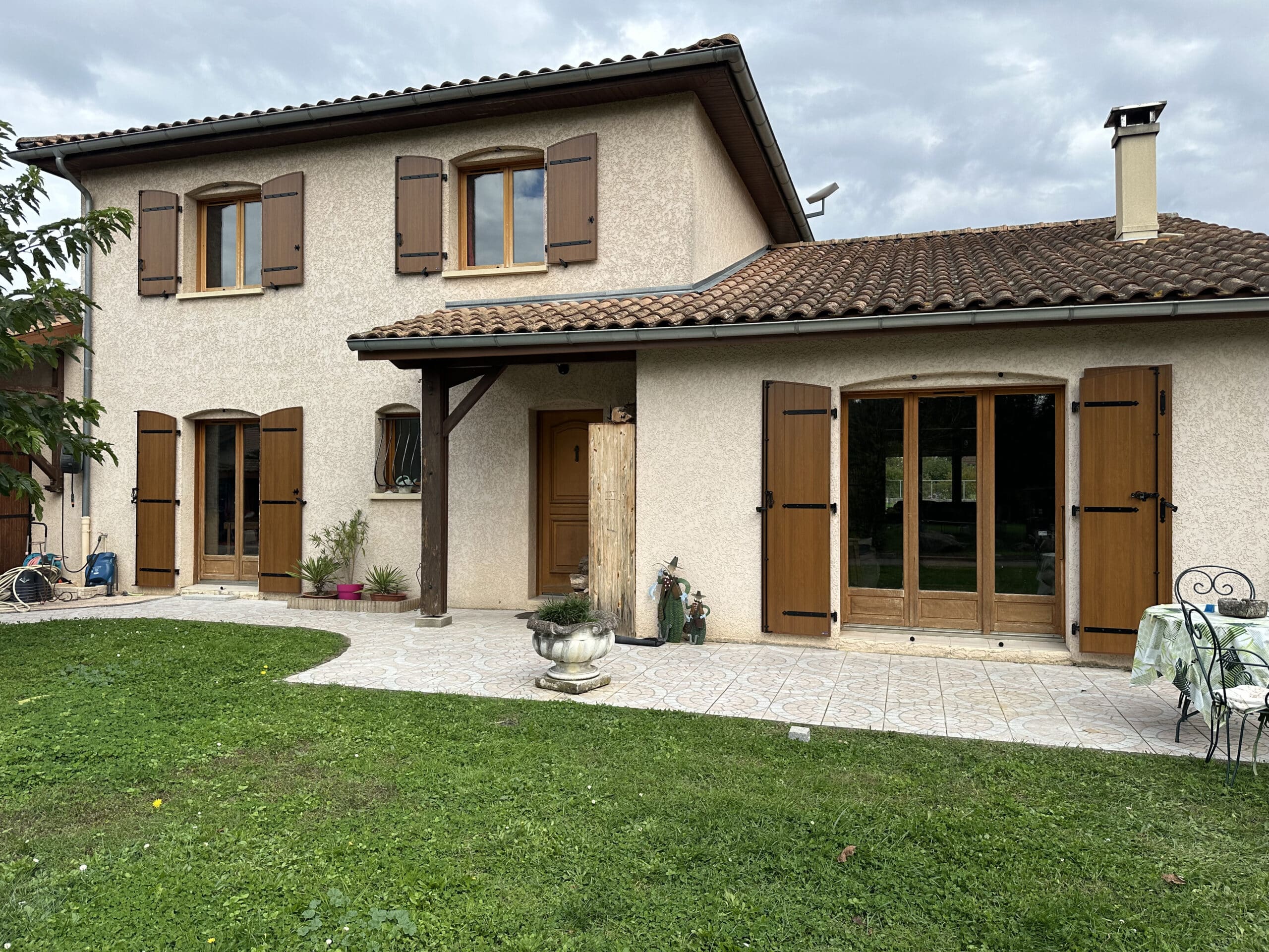 Maison provençale à deux étages avec volets marron, toit en tuiles et terrasse carrelée devant un jardin verdoyant.
