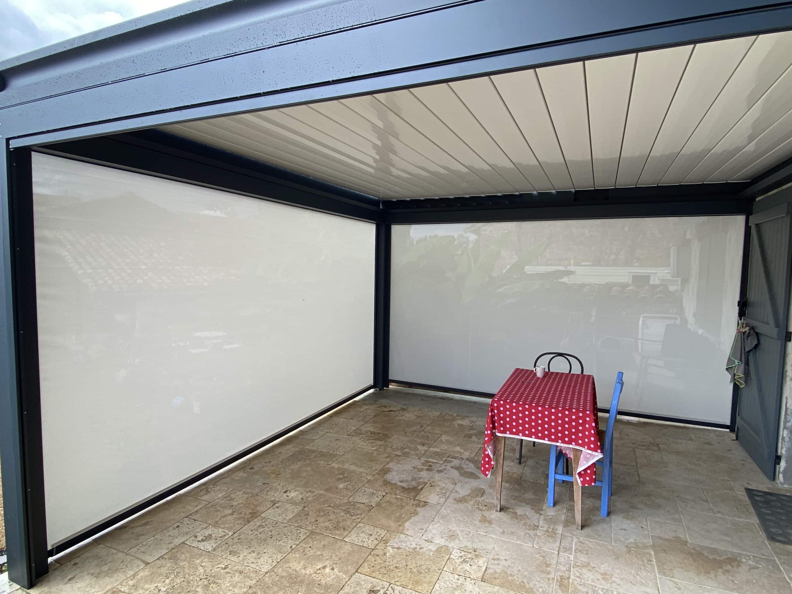 Pergola bioclimatique avec stores latéraux blancs sur une terrasse en pierre avec table de jardin rouge.