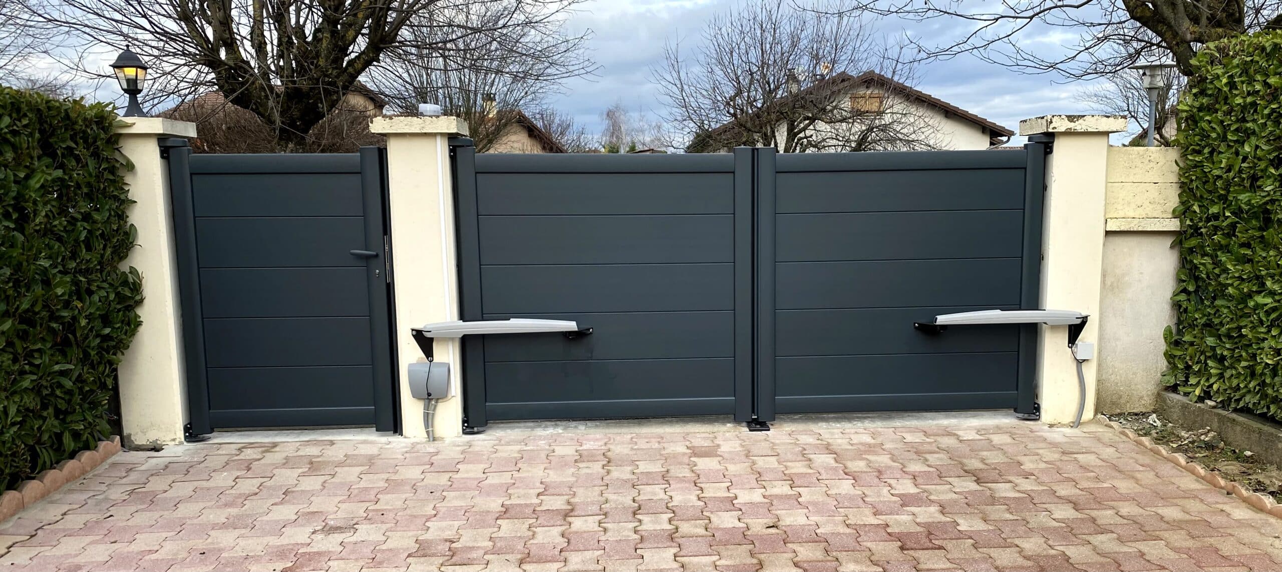 Portail battant et portillon en aluminium gris anthracite motorisés installés entre deux piliers blancs.