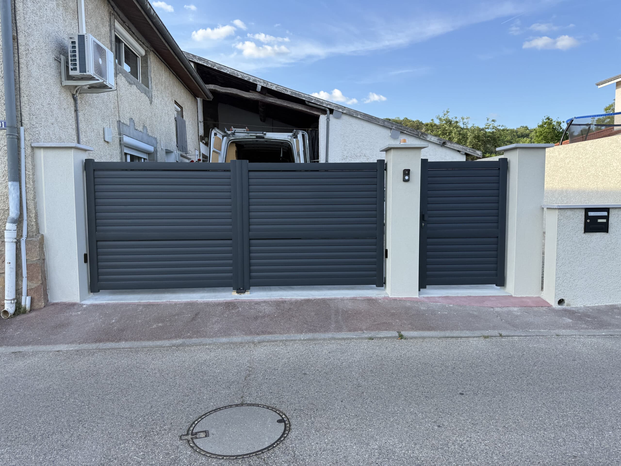 Portail et portillon gris anthracite à lames horizontales installés entre des piliers blancs devant une maison.