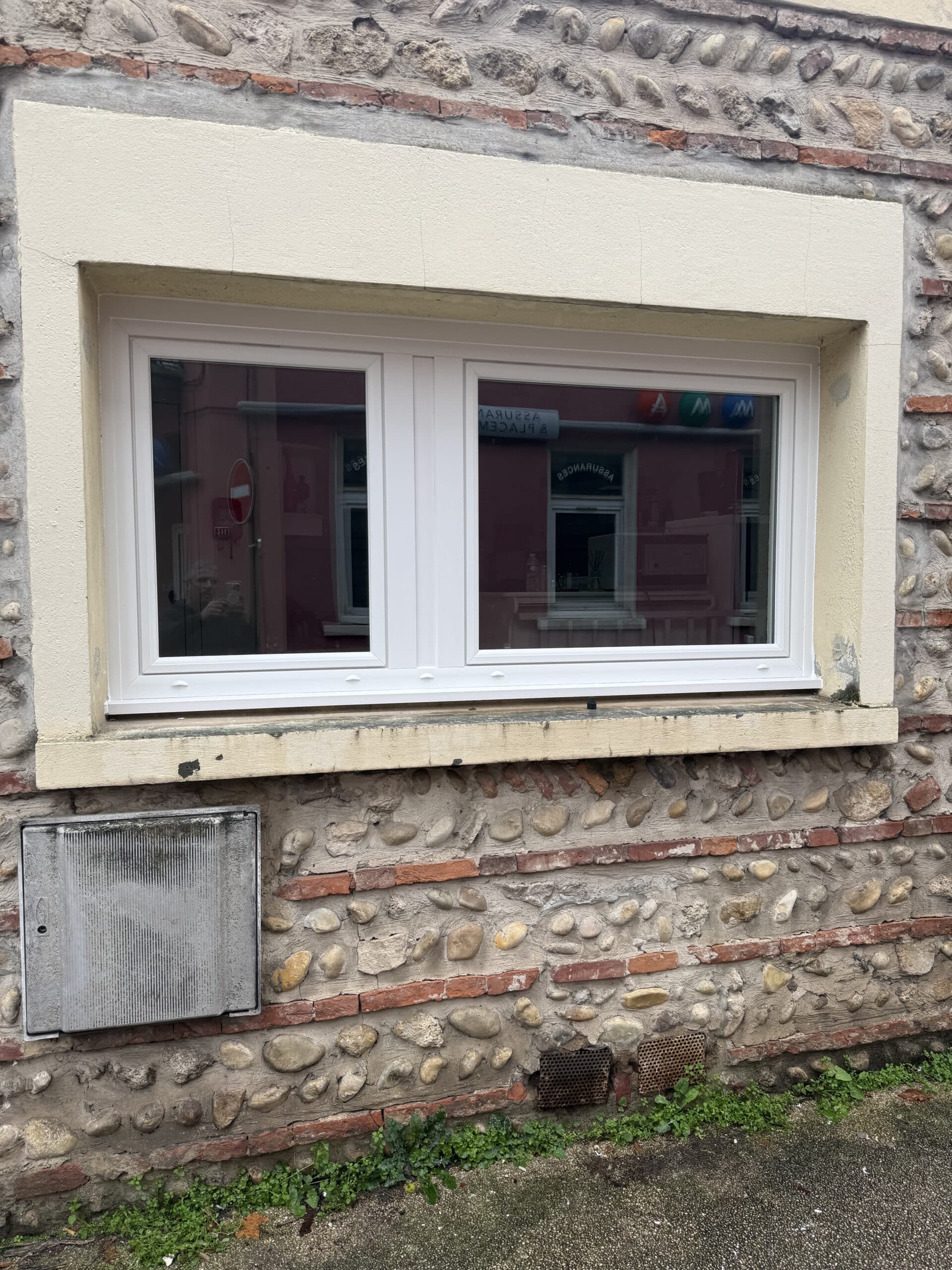 Fenêtre PVC blanche sur mur en pierre et brique, reflet de bâtiment rose avec texte Assurances.